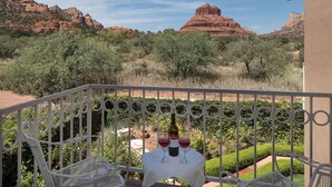 A balcony with a white metal railing, a table with a white tablecloth, and two wine glasses with a bottle of wine. The view includes red rock formations and green vegetation.