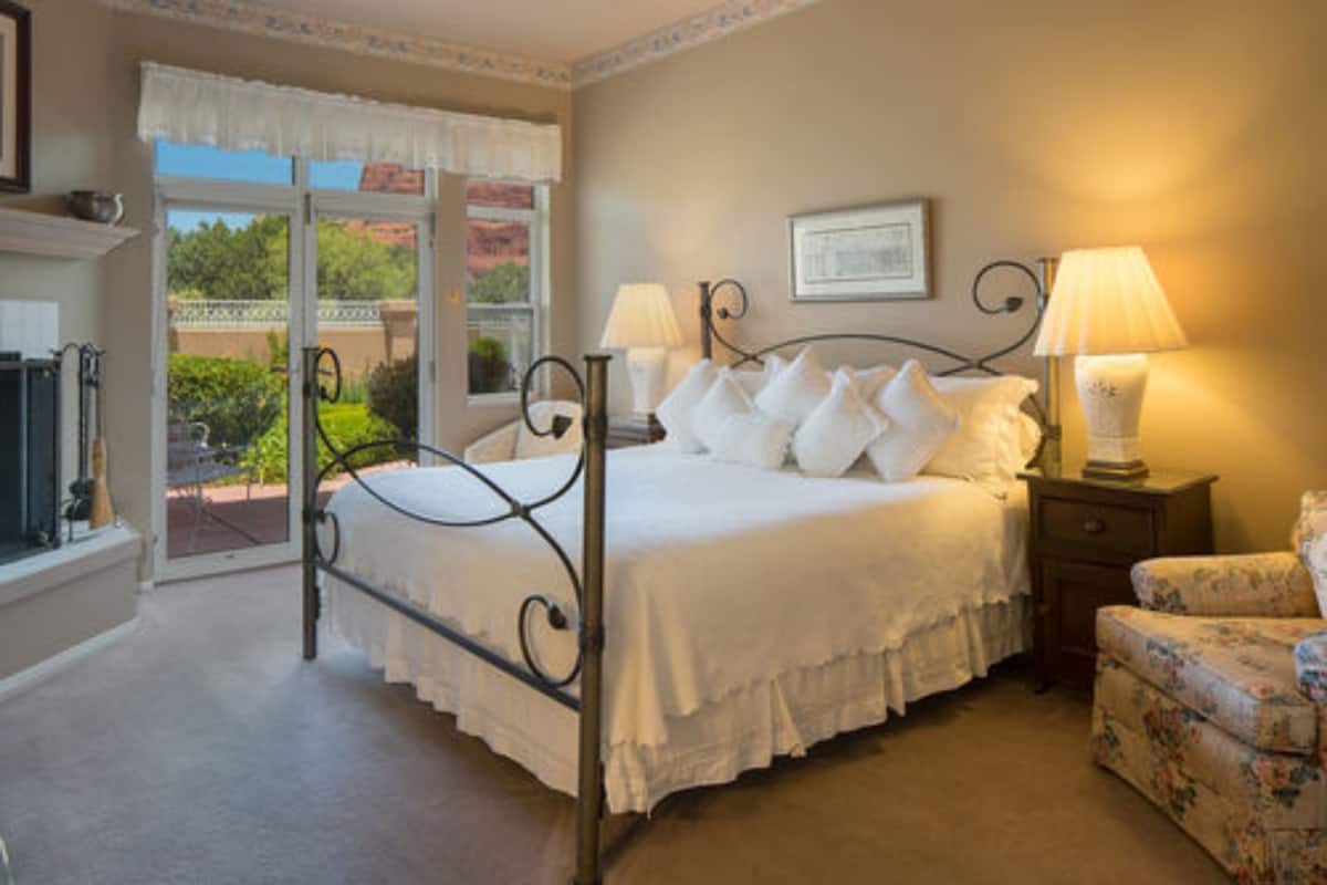 A bedroom with a four-poster bed, two bedside lamps, a floral armchair, and a view of the outdoors through a glass door.