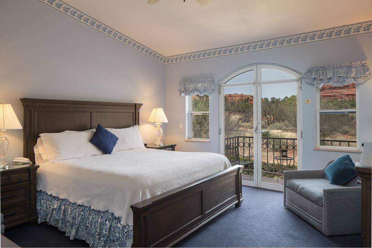 A bedroom with a large bed, two bedside tables with lamps, a nightstand, a window with a view, and a balcony with a chair.