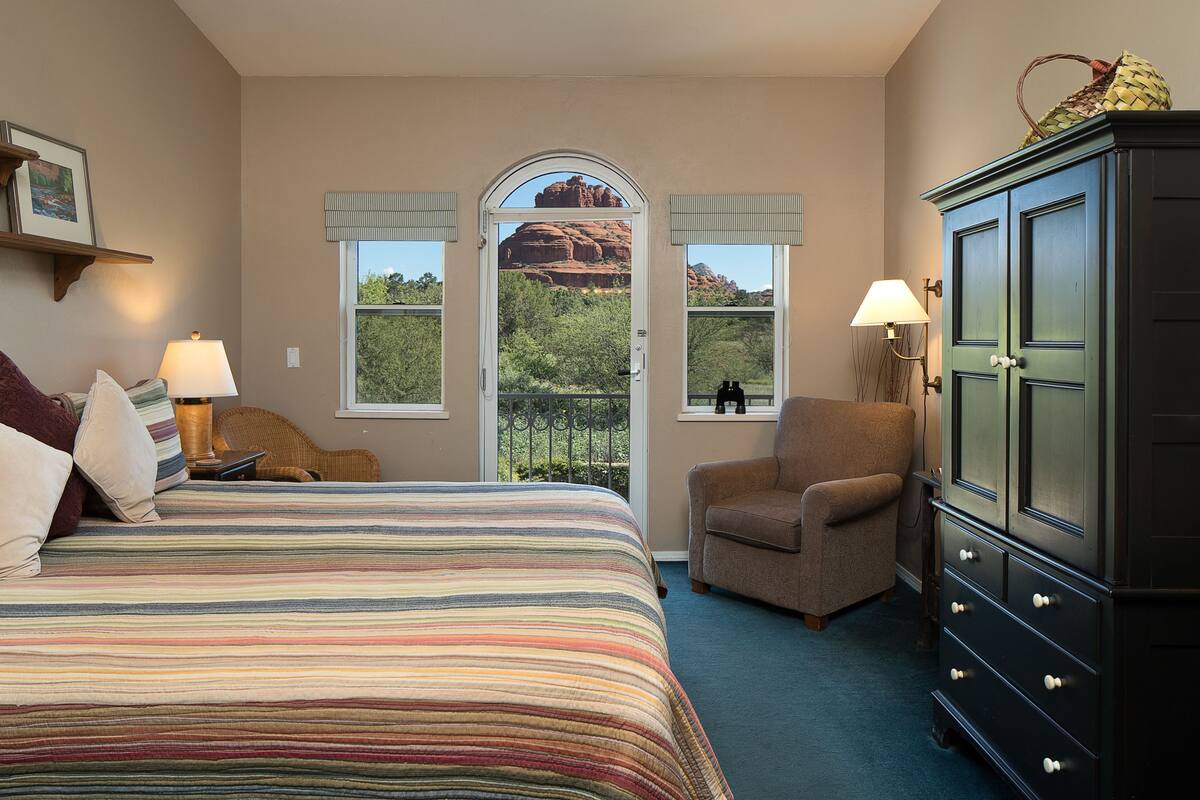 A bedroom with a bed, a chair, a dresser, and a window with a view of red rock formations.