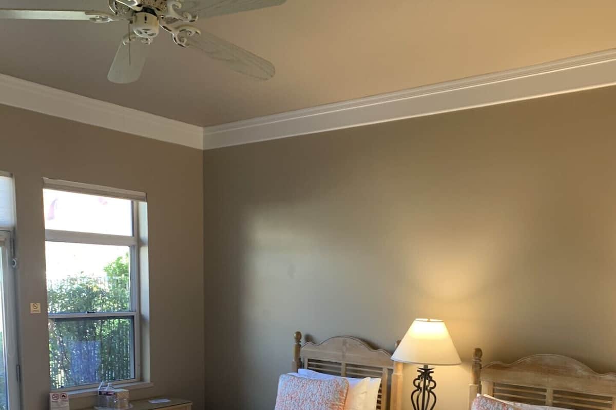 A bedroom with two beds, a ceiling fan, a nightstand with a lamp, and a window with a view of greenery outside.