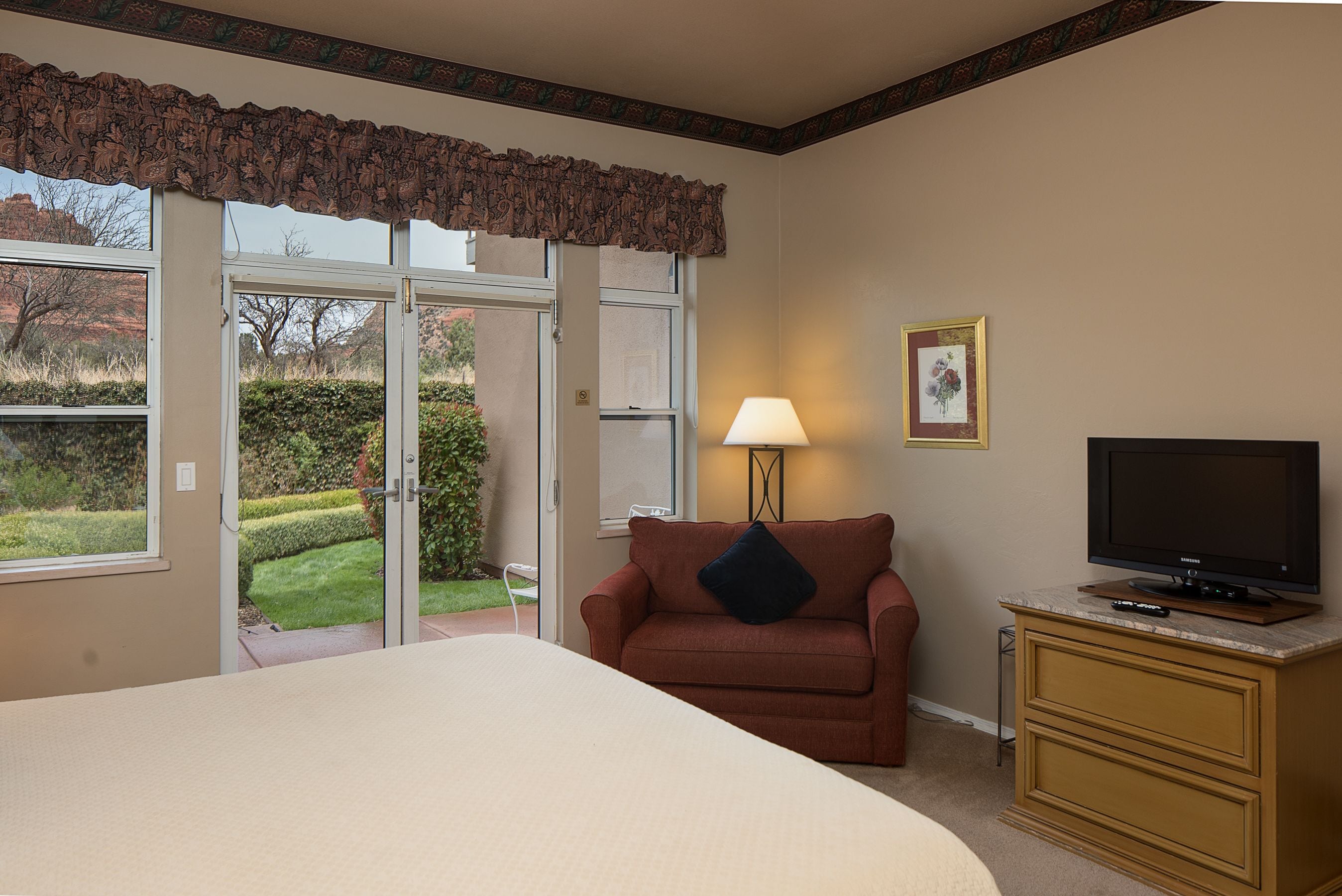 Quarto de hotel com uma cama grande, um sofá marrom, uma televisão sobre um móvel de madeira e uma porta que dá para um jardim.
