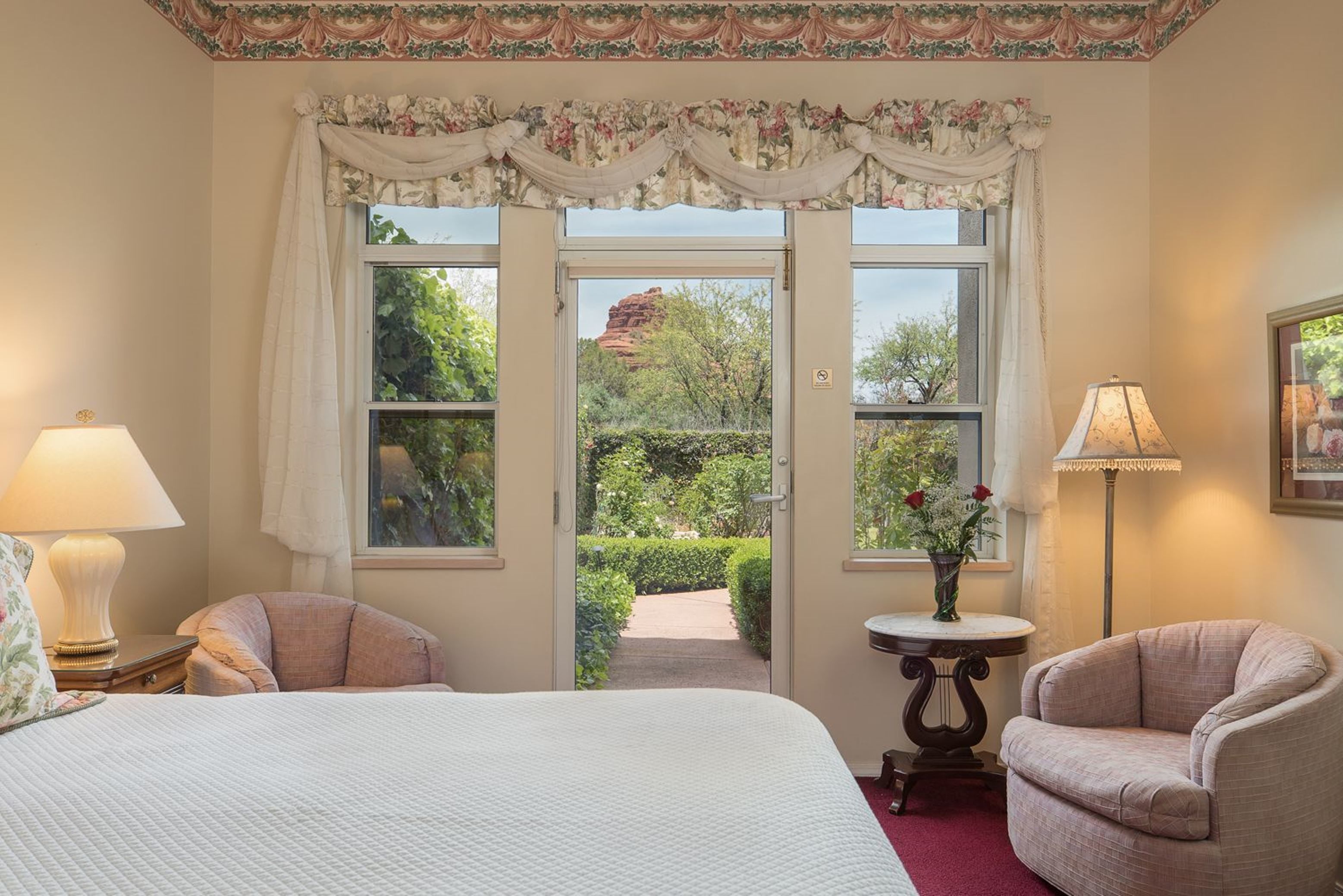 A bedroom with a large bed, two chairs, a table with a vase of flowers, and a view of a garden through a door.