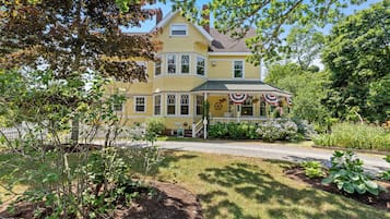 A two-story yellow house with a porch, surrounded by a garden and trees.