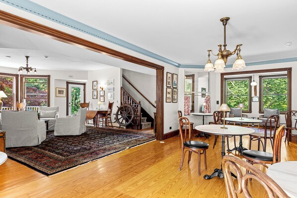 A spacious living room with a large rug, wooden flooring, a staircase, and a dining area with chairs and tables. - The Inn at the Oaks (Eastham)