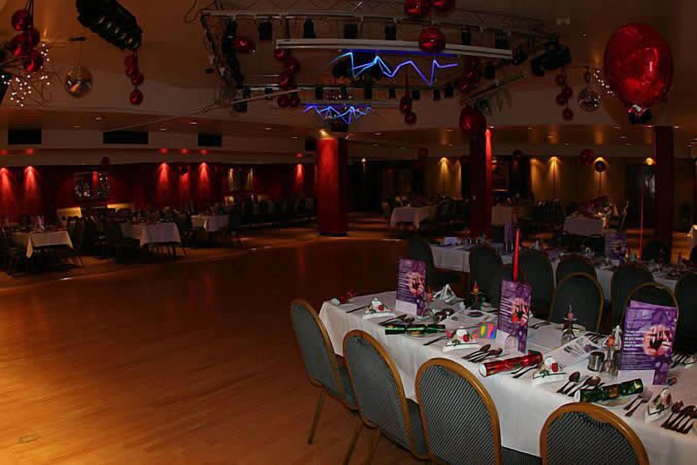 A banquet hall with tables set for a formal event, decorated with red balloons and a neon light fixture.