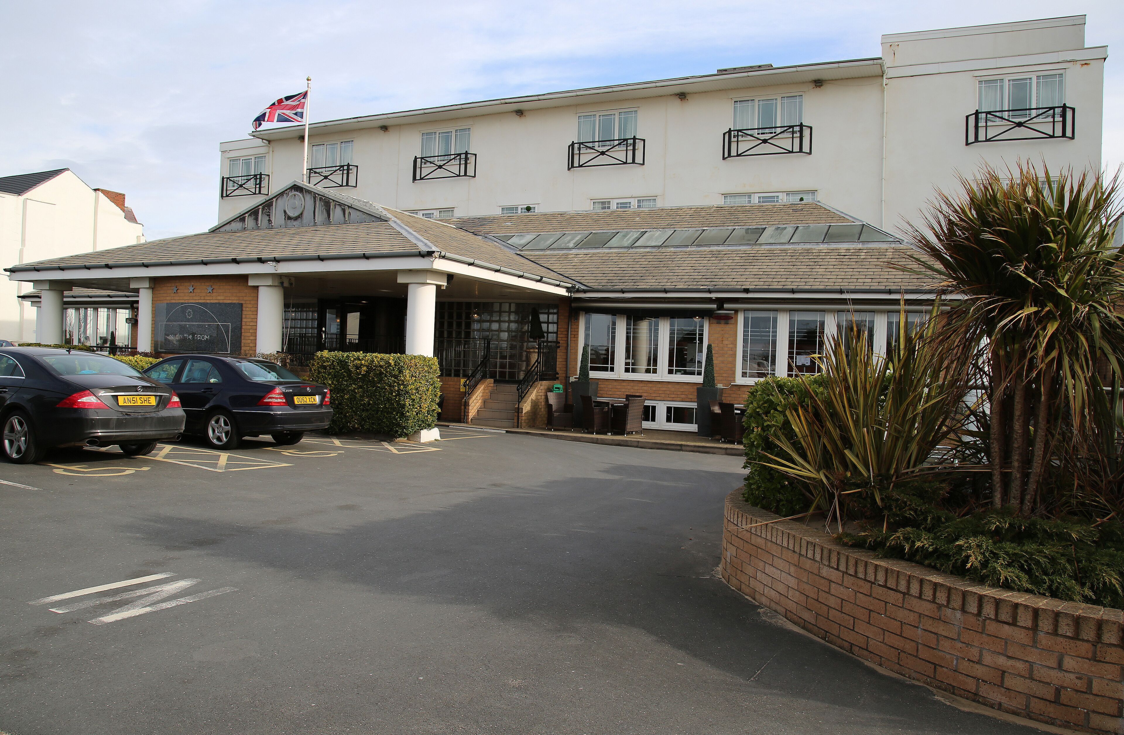 A hotel with a covered entrance, a flag on top, and a parking lot with two cars.