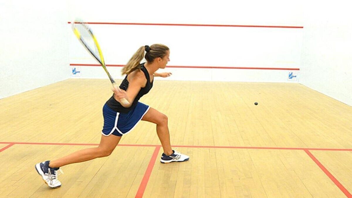 Una cancha de squash con un jugador en movimiento, piso de madera y líneas divisorias rojas.