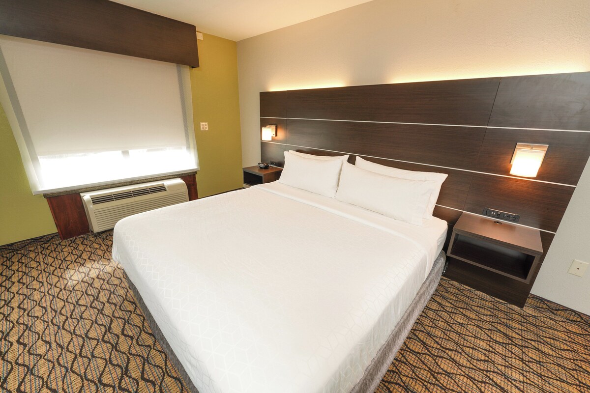 A hotel room with a large bed, a nightstand, a wall-mounted lamp, and a window with blinds.