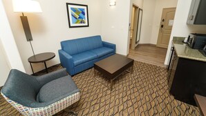 A living room with a blue sofa, a small round table, a patterned carpet, and a kitchenette area. - Holiday Inn Express and Suites Springfield Medical District by IHG (Springfield)