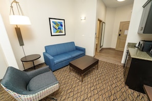 Ein Wohnzimmer mit einem blauen Sofa, einem kleinen runden Tisch, einem gemusterten Teppich und einer Küchenzeile.
