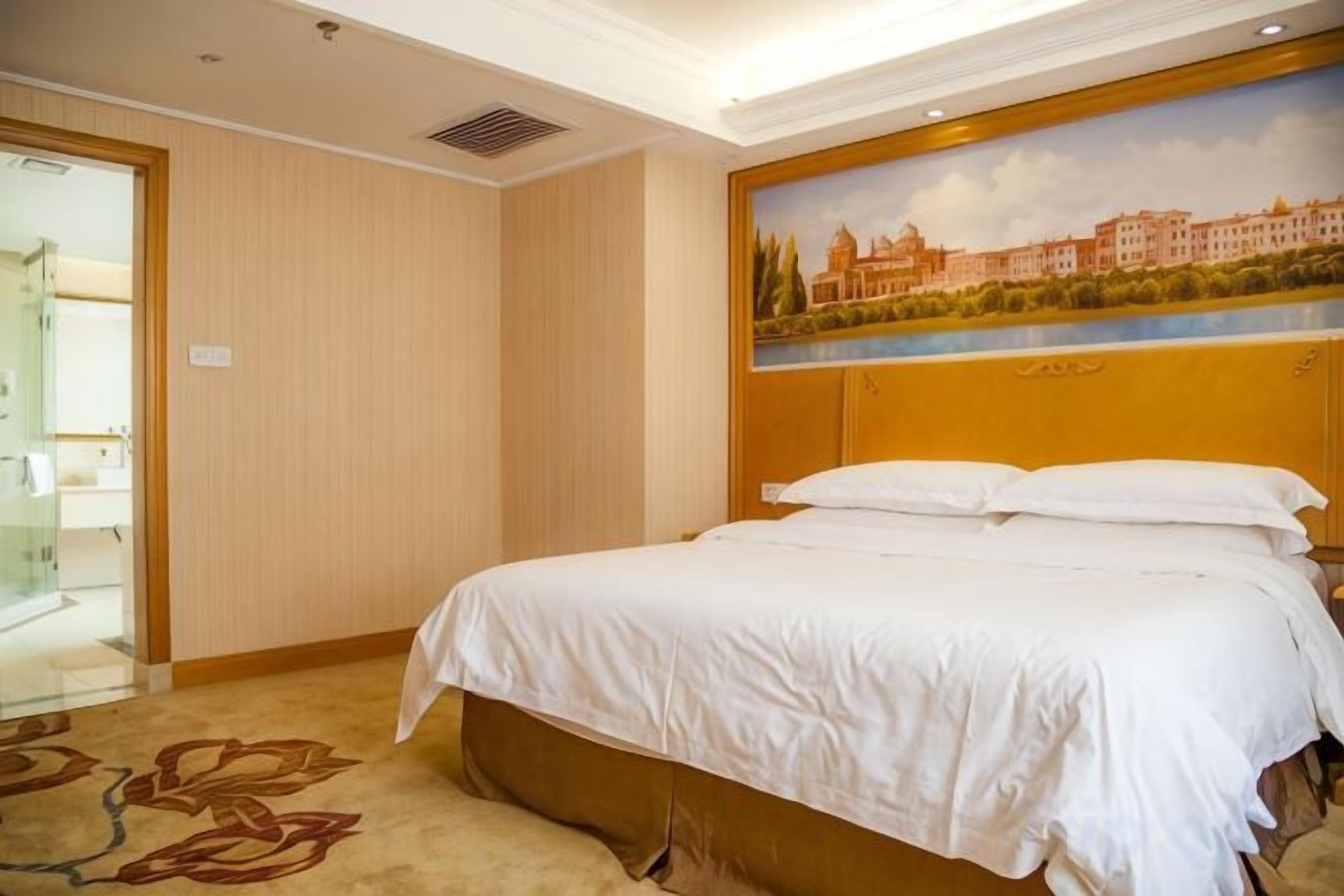 A hotel room with a bed, a painting of a cityscape, and a door leading to a bathroom.