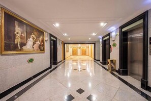 A hotel corridor with a large classical painting on the wall, a carpeted floor, and a reception area in the distance. - Vienna International Hotel (Beijing Niujie Guang'anmennei Subway Station Branch) (Beijing)