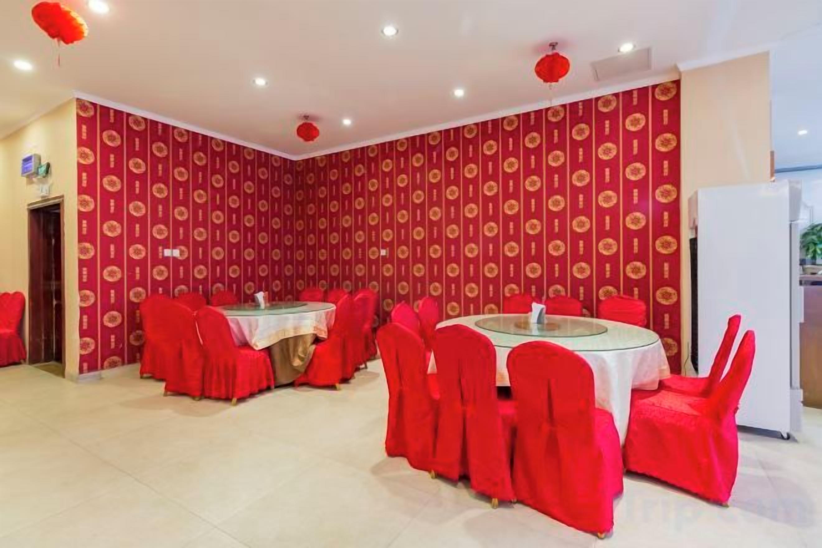 Un espace repas avec des tables rondes et des chaises rouges, un mur orné de motifs et un réfrigérateur blanc.