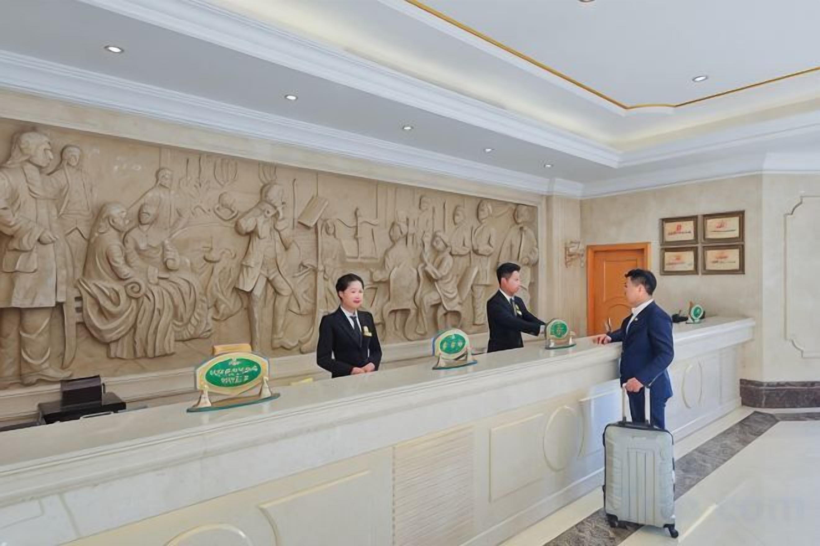 A reception desk with three staff members, a marble wall with relief sculptures, and a wooden door.