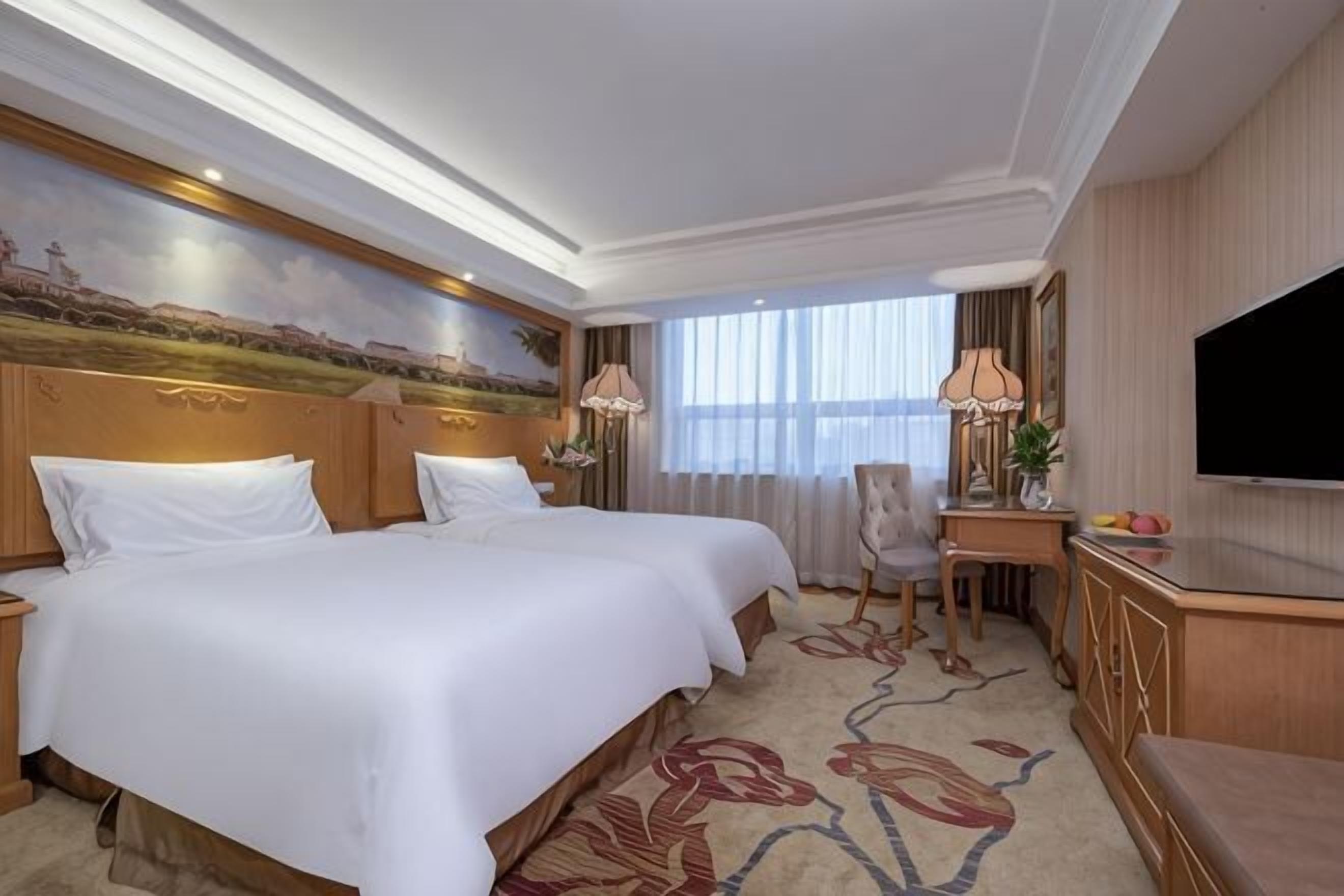 Habitación de hotel con dos camas, un escritorio, un televisor y un cuadro en la pared.