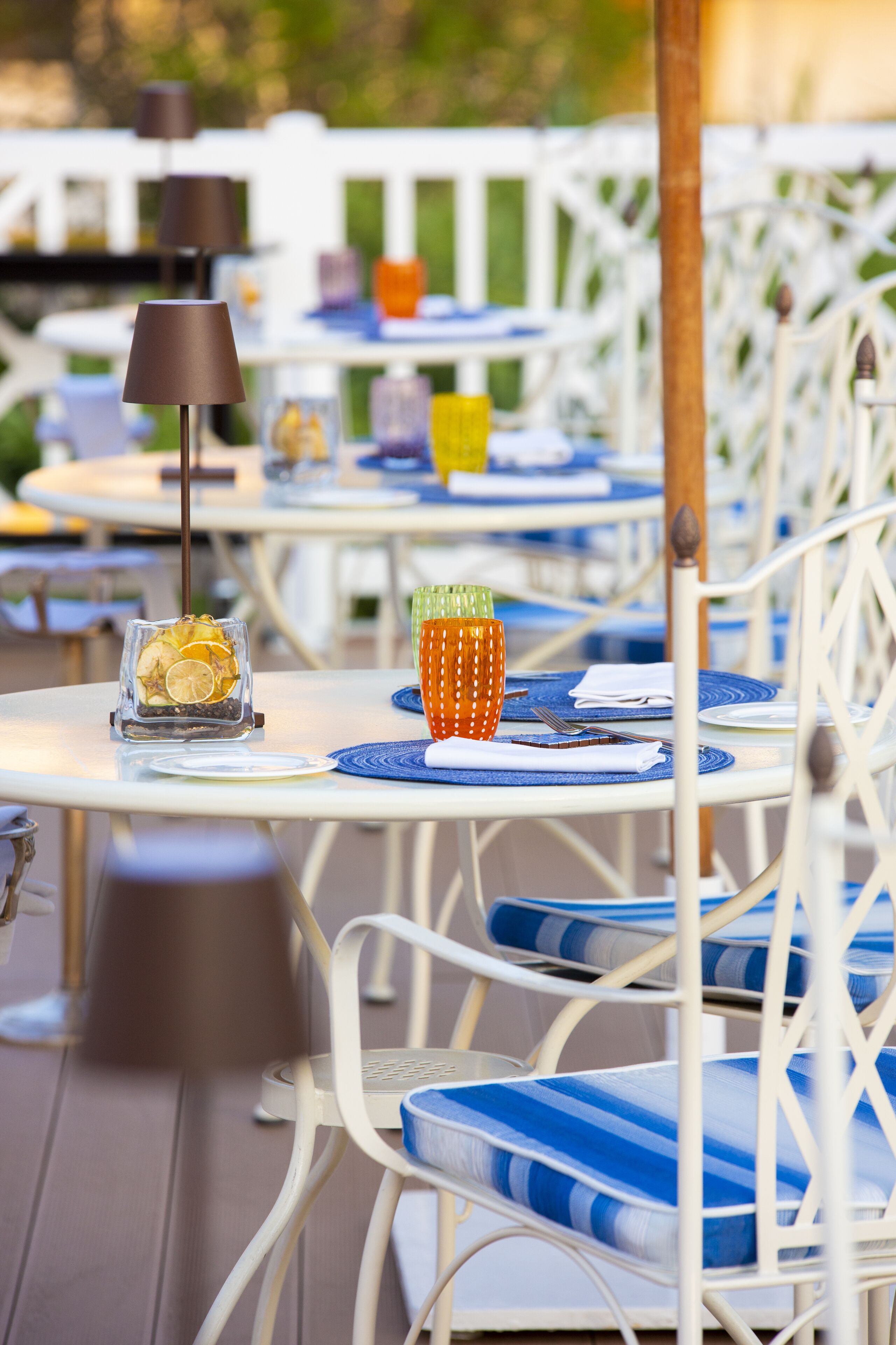 Outdoor dining tables with chairs, each set with a placemat, a glass container with a lemon slice, and a candle holder.