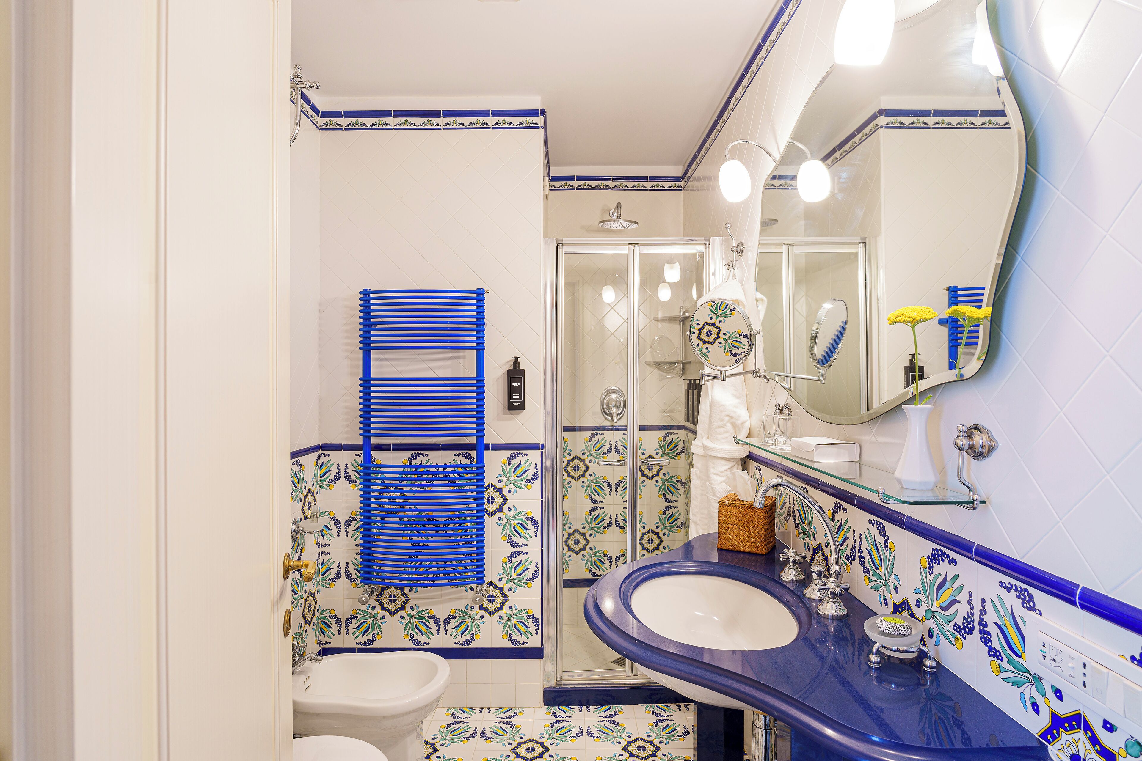 Une salle de bain avec un sol carrelé de blanc et de bleu, un porte-serviettes bleu, une cabine de douche en verre et un lavabo avec un miroir au-dessus.