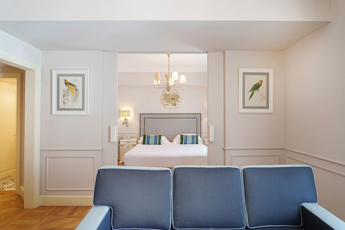 Een hotelkamer met een groot bed, twee blauwe stoelen, een kroonluchter en kunstwerken in lijsten.