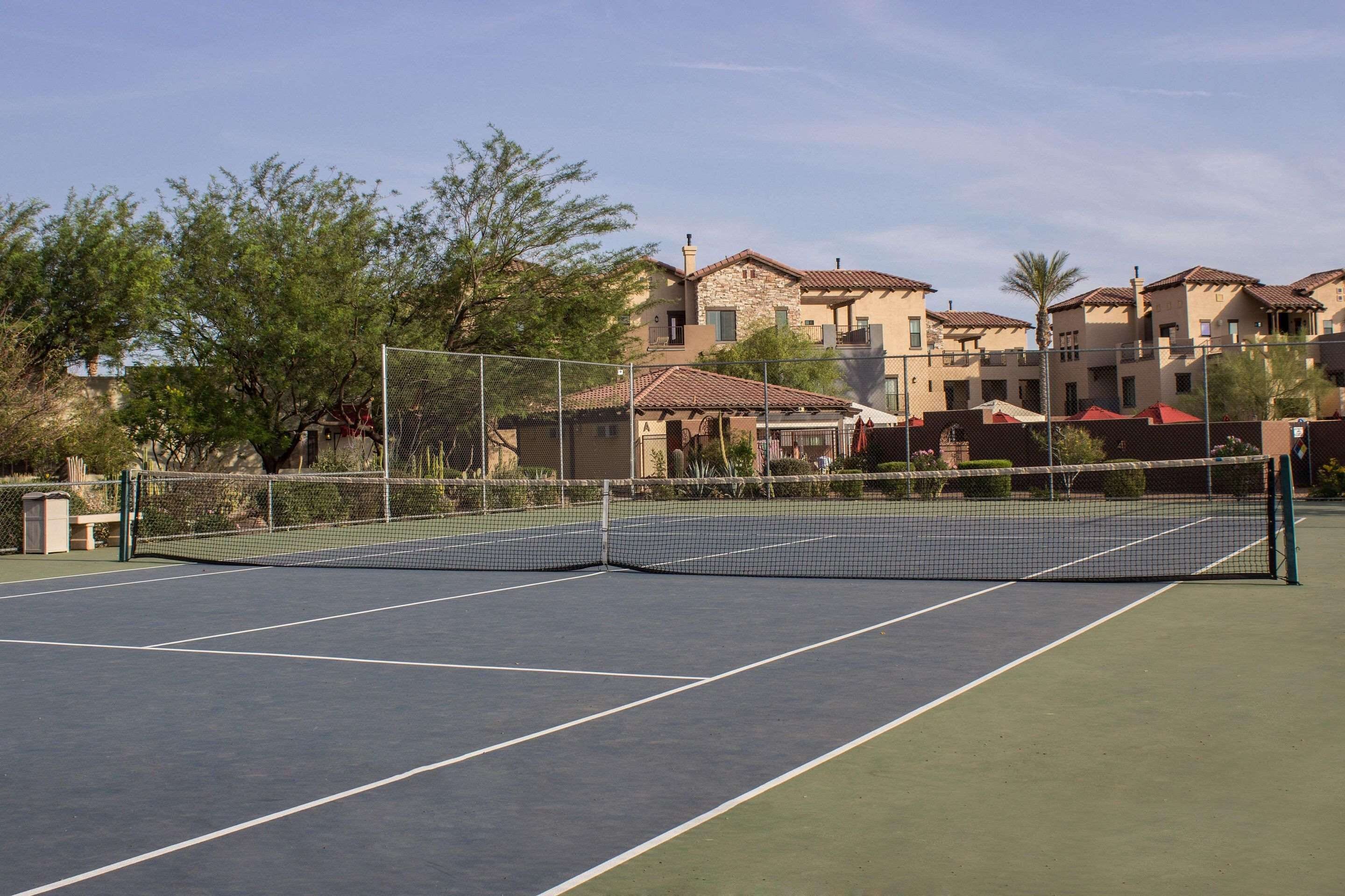 An outdoor tennis court with a net, surrounded by a fence.