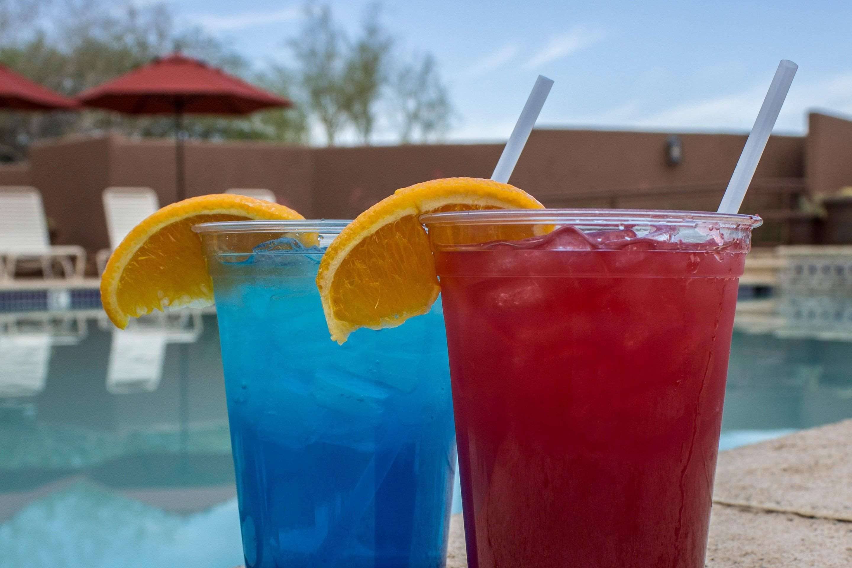 Two plastic cups with straws and orange slices, placed by a poolside with umbrellas in the background.