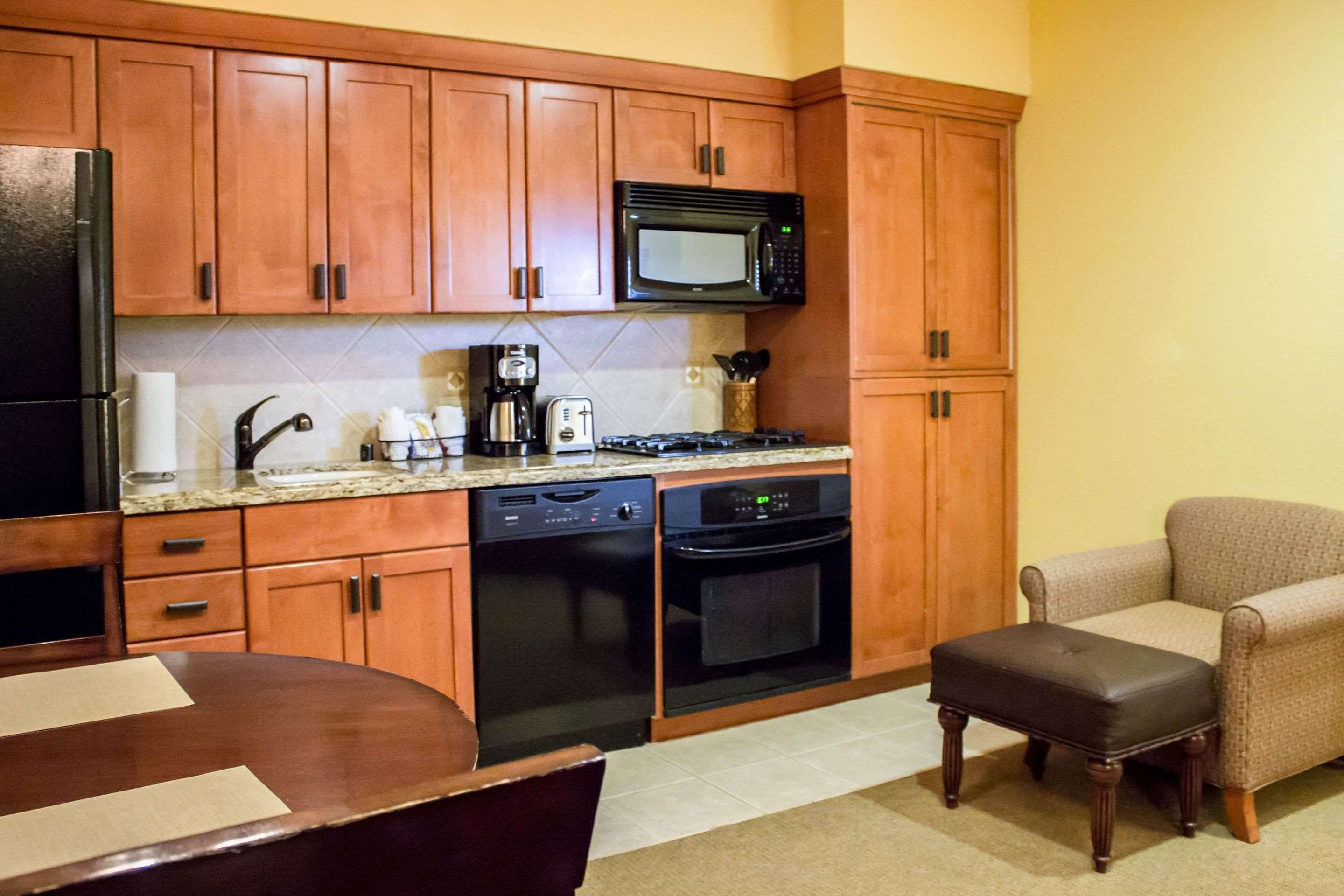 A compact kitchen with wooden cabinets, a microwave, a dishwasher, and a stove. There is a dining table with chairs and a small ottoman.