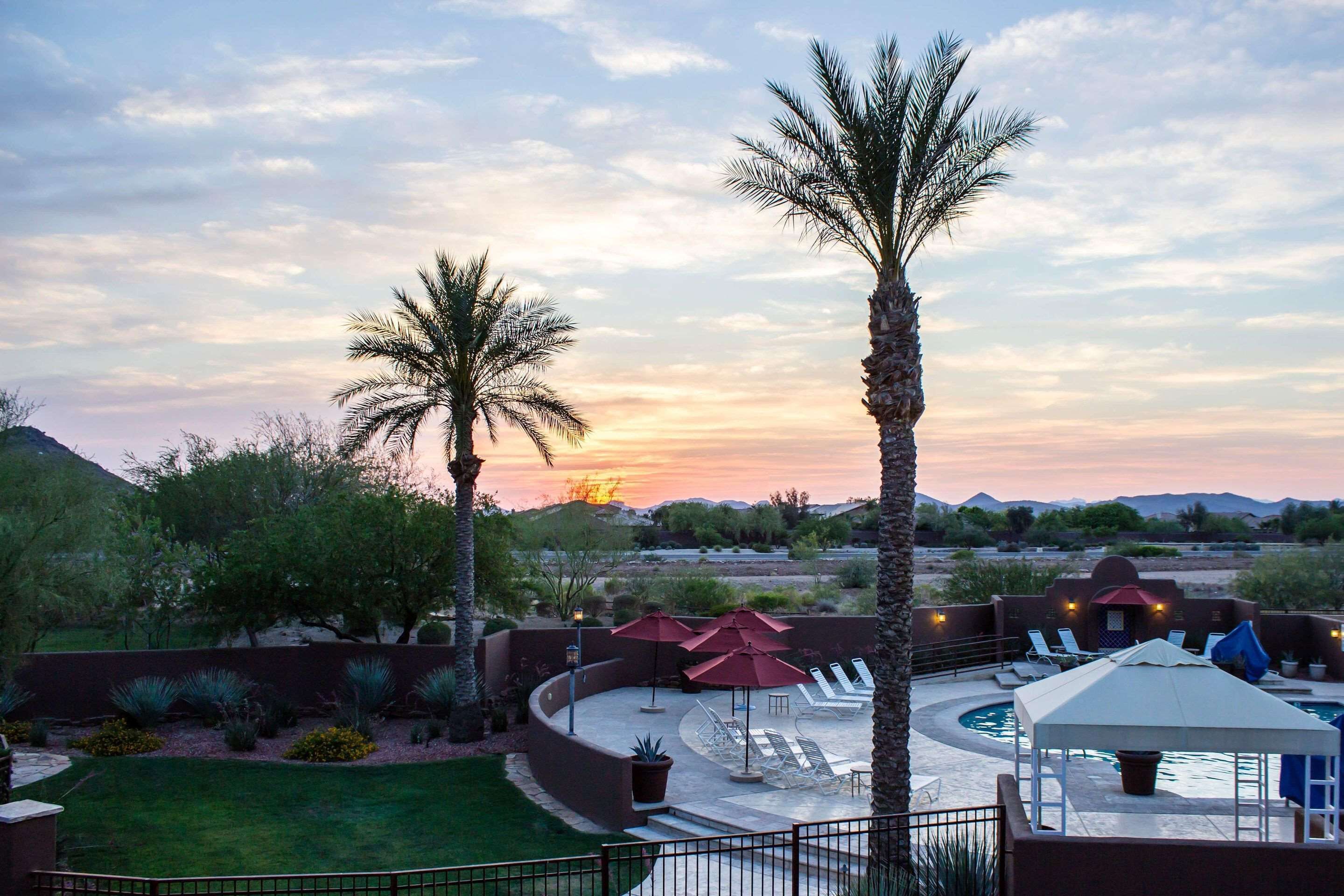 A pool area with lounge chairs and umbrellas, surrounded by palm trees and a sunset view.