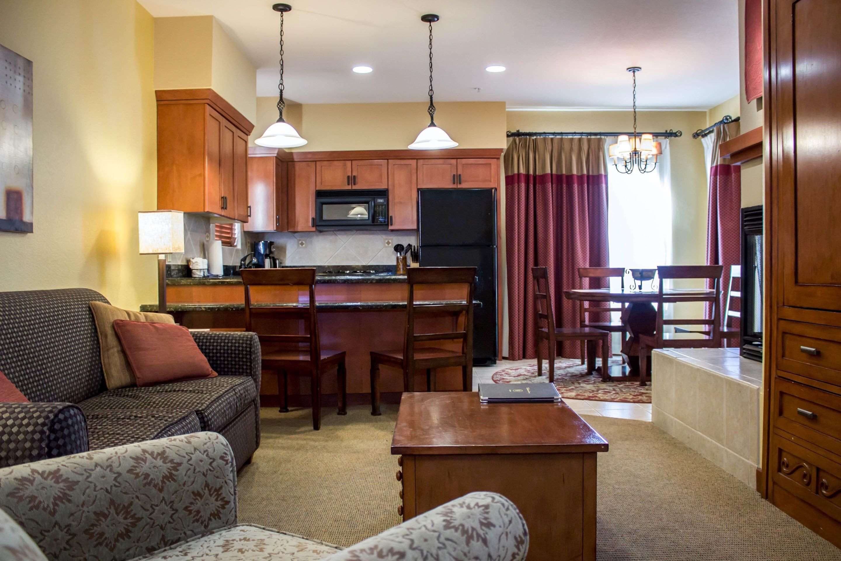 A cozy living room with a sofa, a coffee table, and a dining area with chairs and a table. A kitchen with wooden cabinets and appliances is visible in the background.