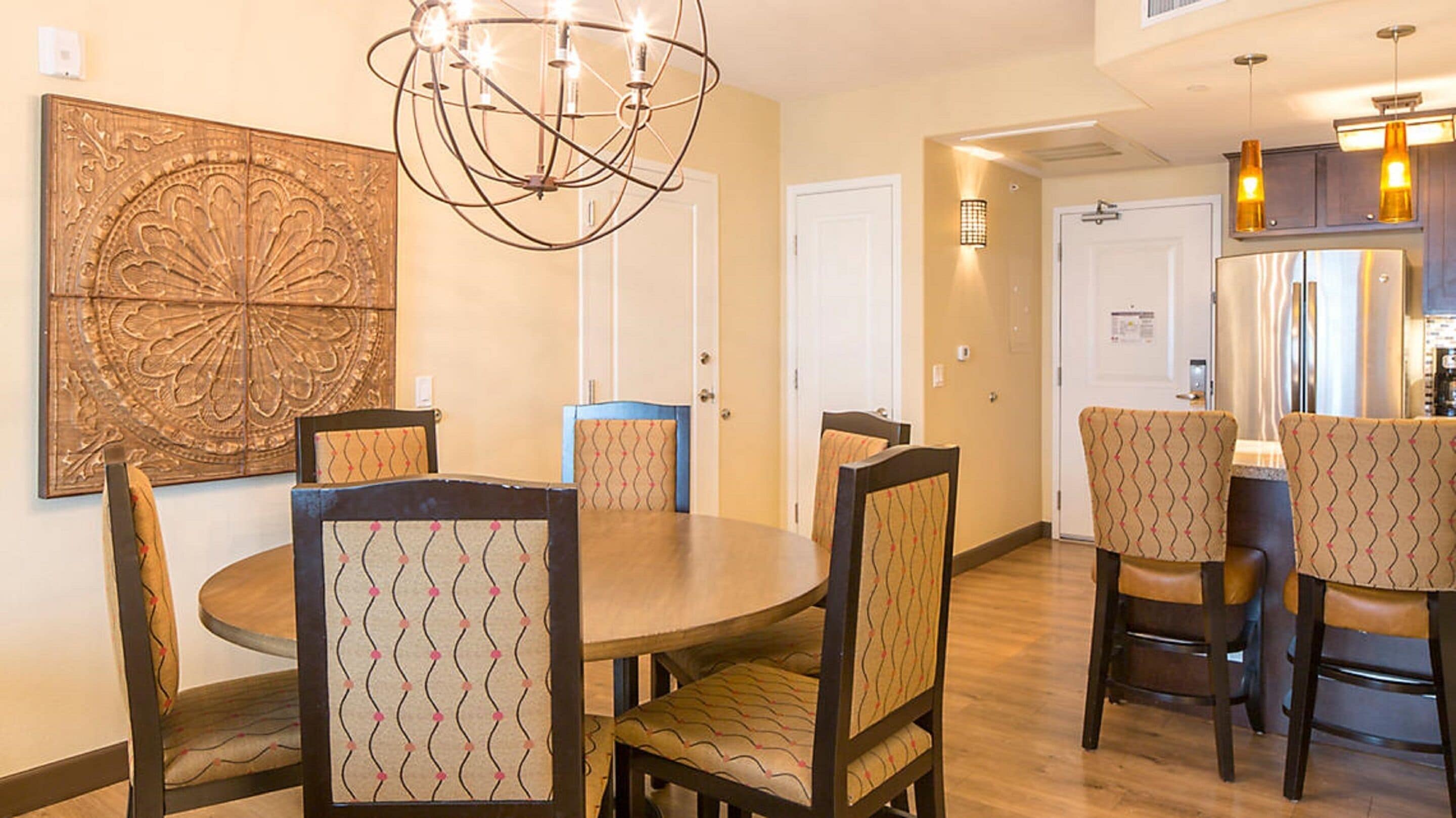 A dining area with a round table and six chairs, a chandelier, and a large decorative wall piece.