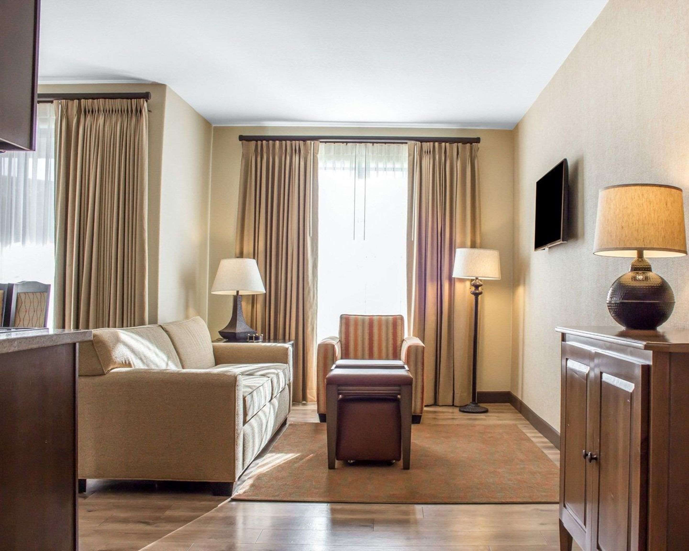 Une chambre d’hôtel équipée d’un canapé, d’un fauteuil, d’un pouf, d’une lampe sur pied et d’un téléviseur fixé au mur.