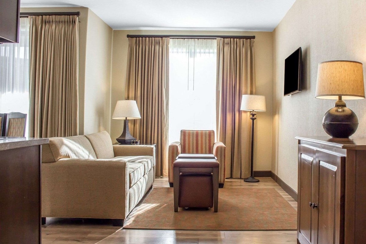 A hotel room with a sofa, armchair, ottoman, floor lamp, and a TV on the wall.