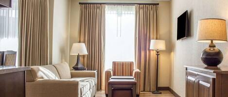 Une chambre d’hôtel équipée d’un canapé, d’un fauteuil, d’un pouf, d’une lampe sur pied et d’un téléviseur fixé au mur.