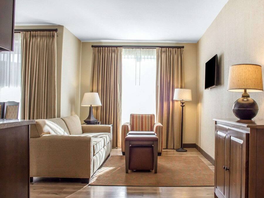 Habitación de hotel con sofá, sillón, reposapiés, lámpara de pie y televisión en la pared.