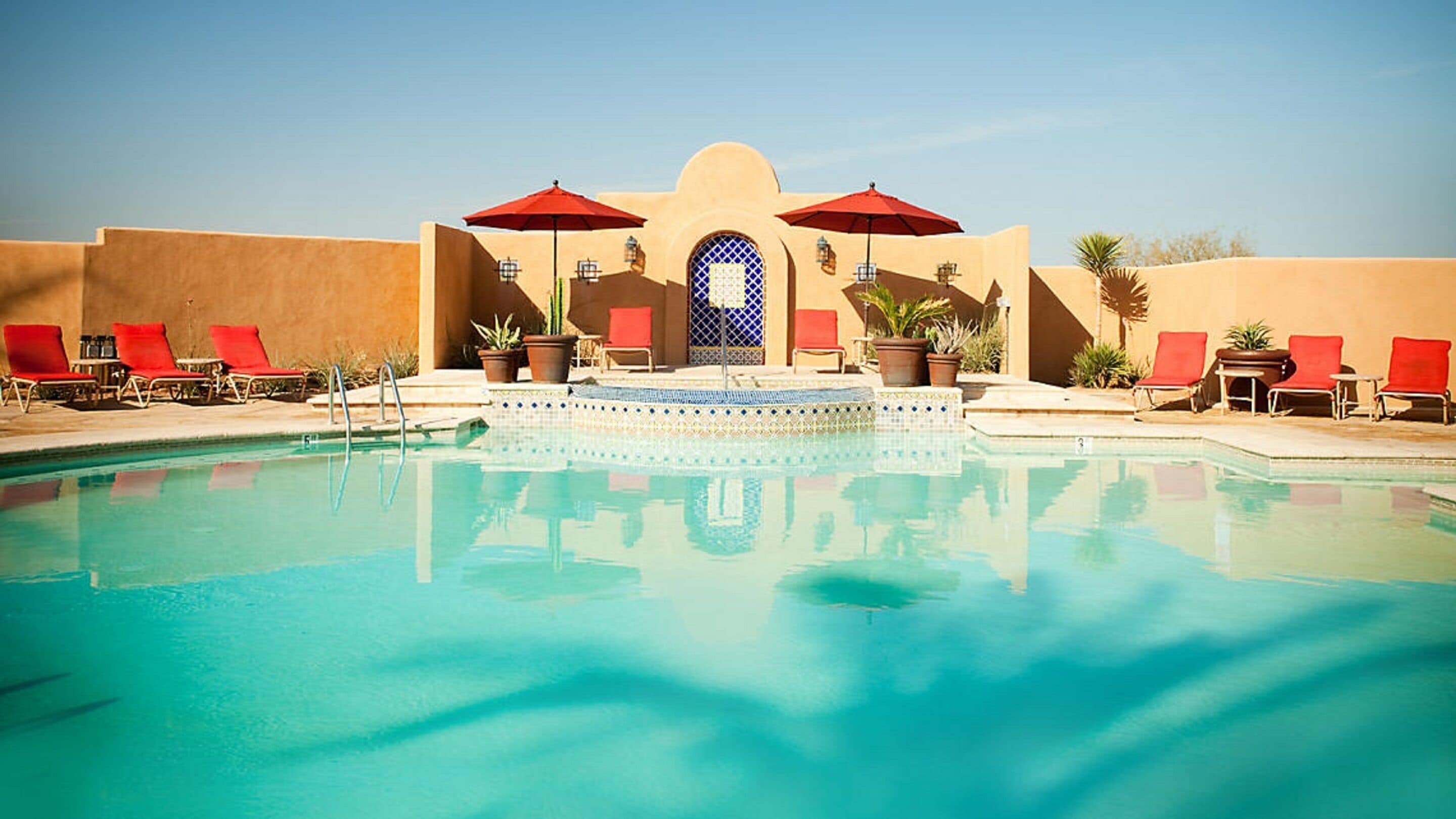 A pool area with a clear blue pool, red umbrellas, and lounge chairs.