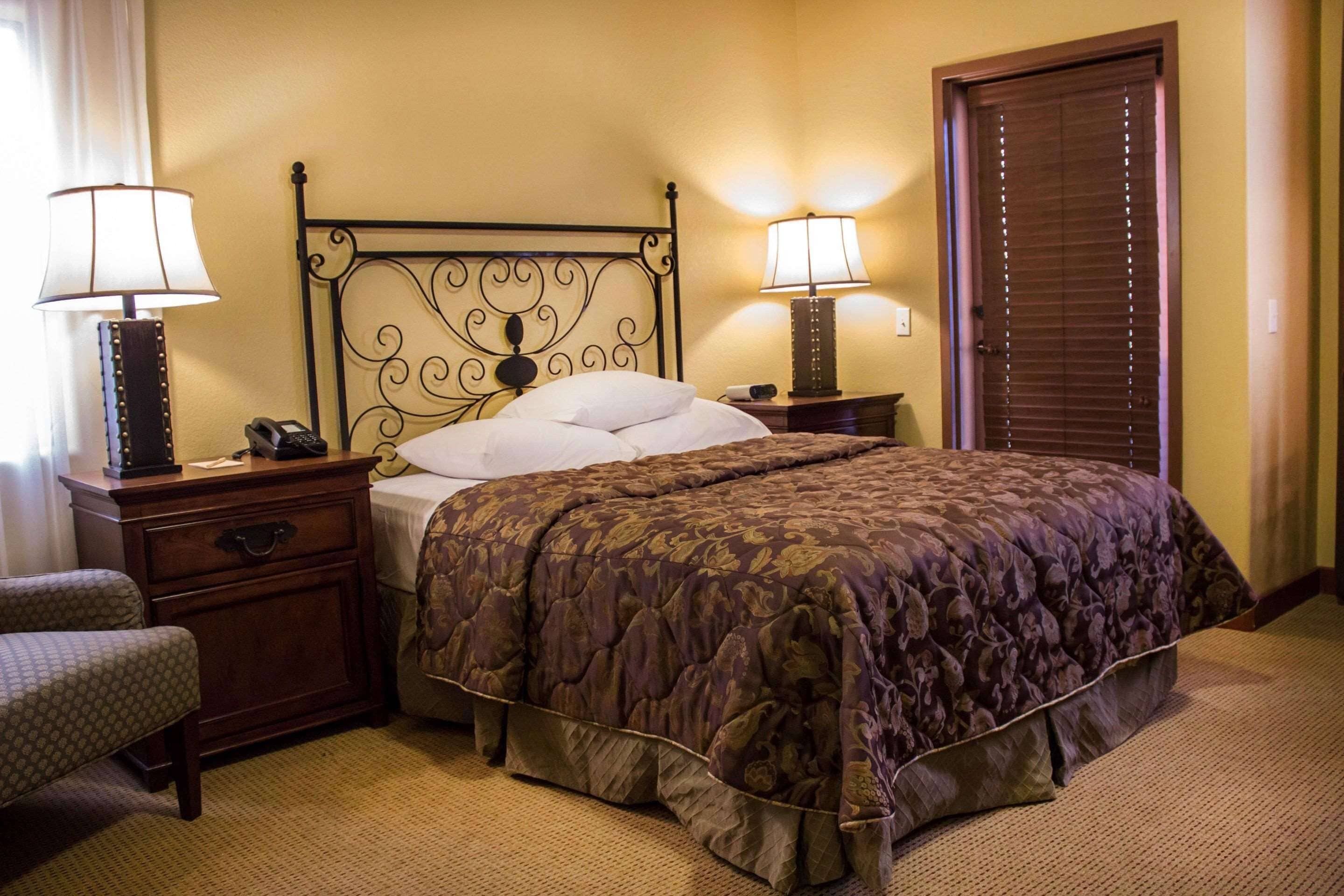 A hotel room with a bed, two bedside tables with lamps, a wooden nightstand, a chair, and a door.