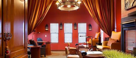 A cozy attic room with a red wall, a chandelier, and a fireplace. There are two windows with blinds, a desk, and a chair.