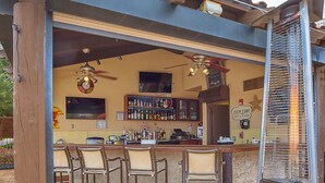 Eine Außenbar mit einer Holzkonstruktion, Barhocker, einem Deckenventilator und einer Auswahl an Flaschen in Regalen.