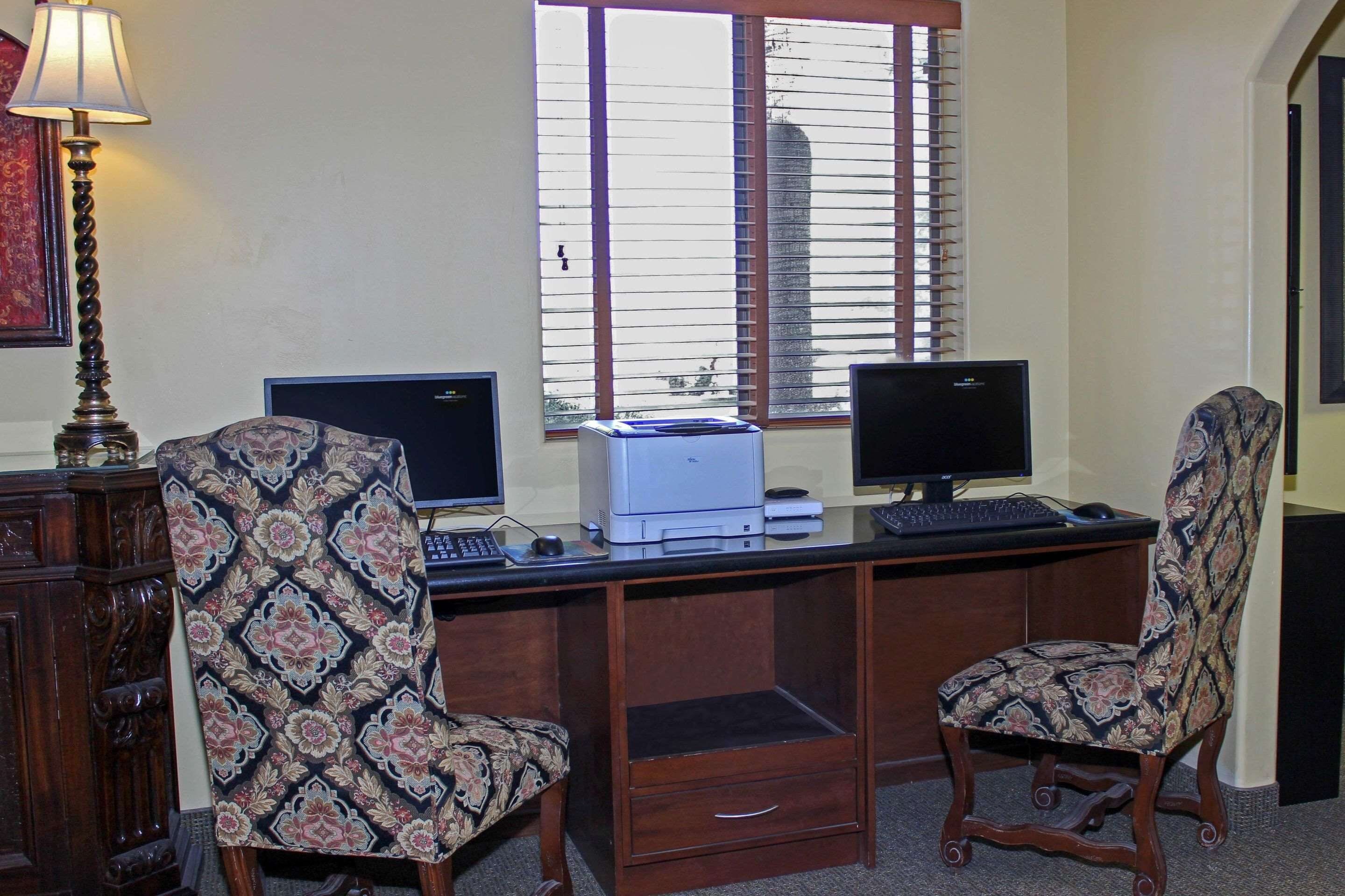 A wooden desk with two monitors, a printer, and patterned chairs in a room with a window and a lamp.
