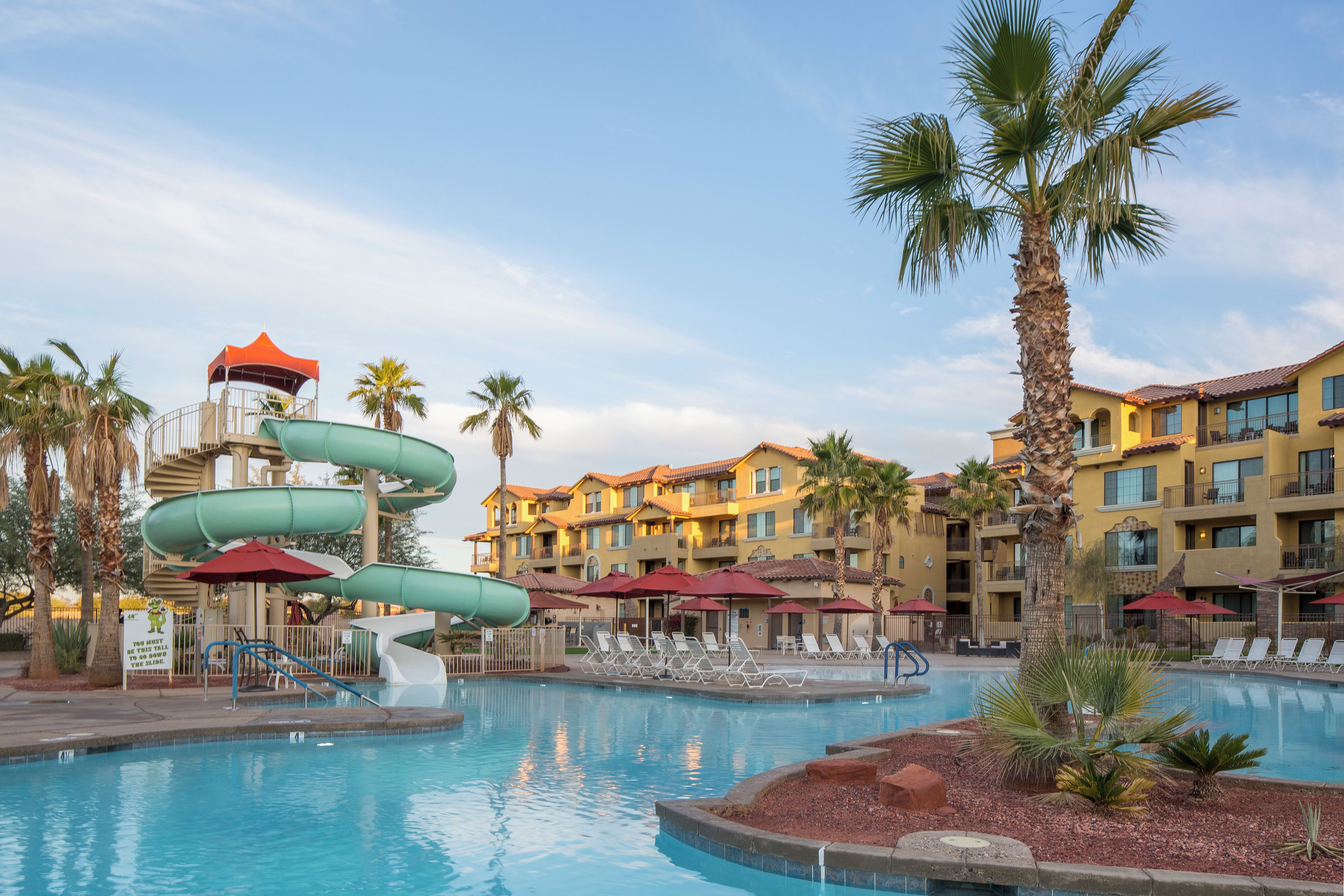 A swimming pool with a water slide, palm trees, and a hotel building with balconies.