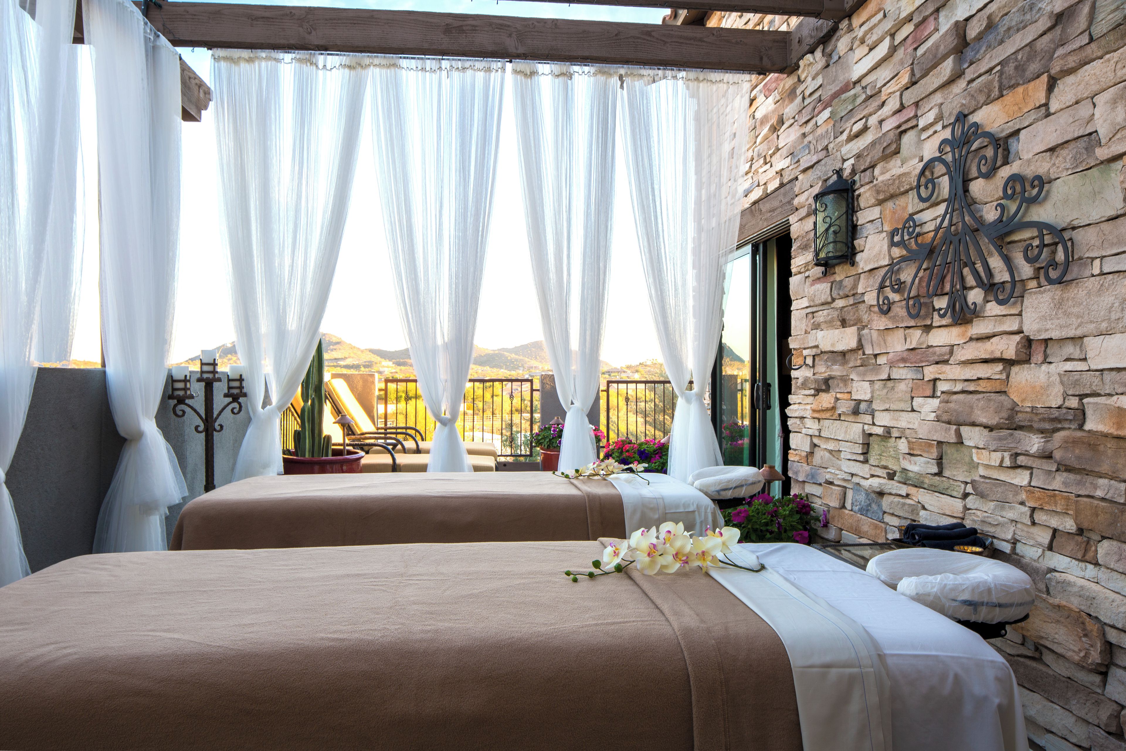 A room with two beds, white curtains, a stone wall, and a view of the mountains.
