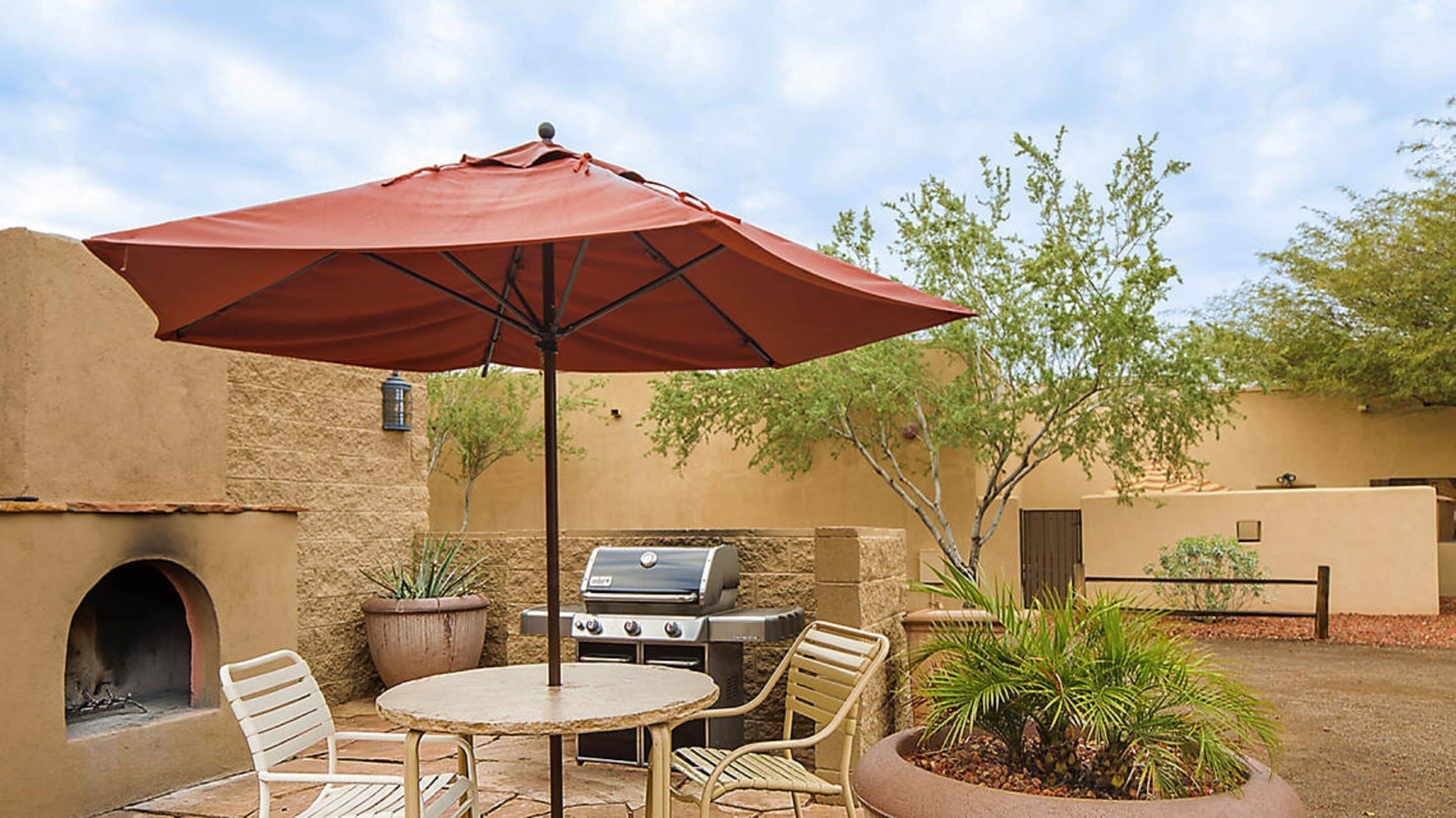 An outdoor patio with a round table, two chairs, a red umbrella, a barbecue grill, and potted plants.
