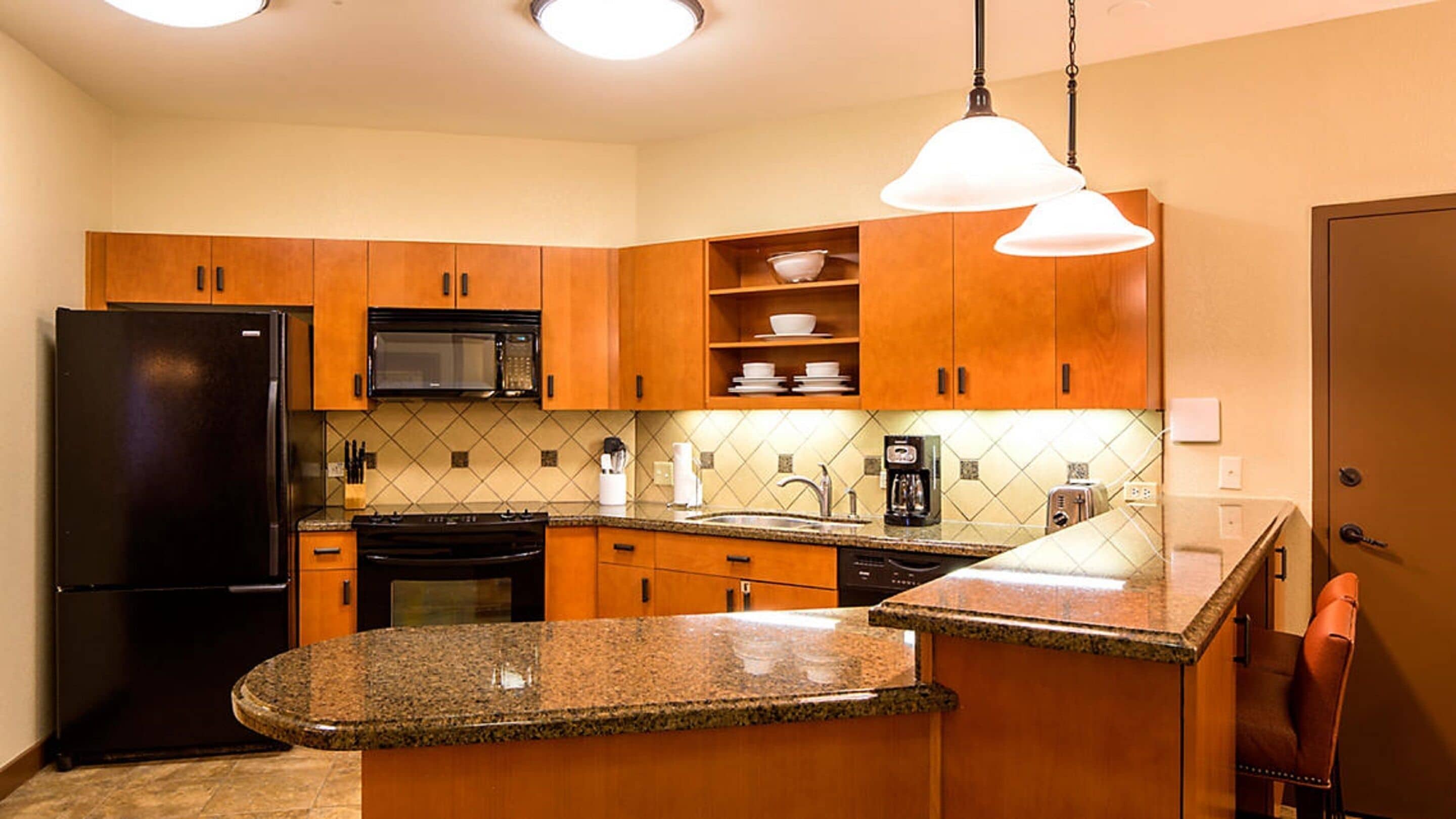 A modern kitchen with wooden cabinets, a black refrigerator, a microwave, a stove, and a sink. There are two pendant lights above the counter.