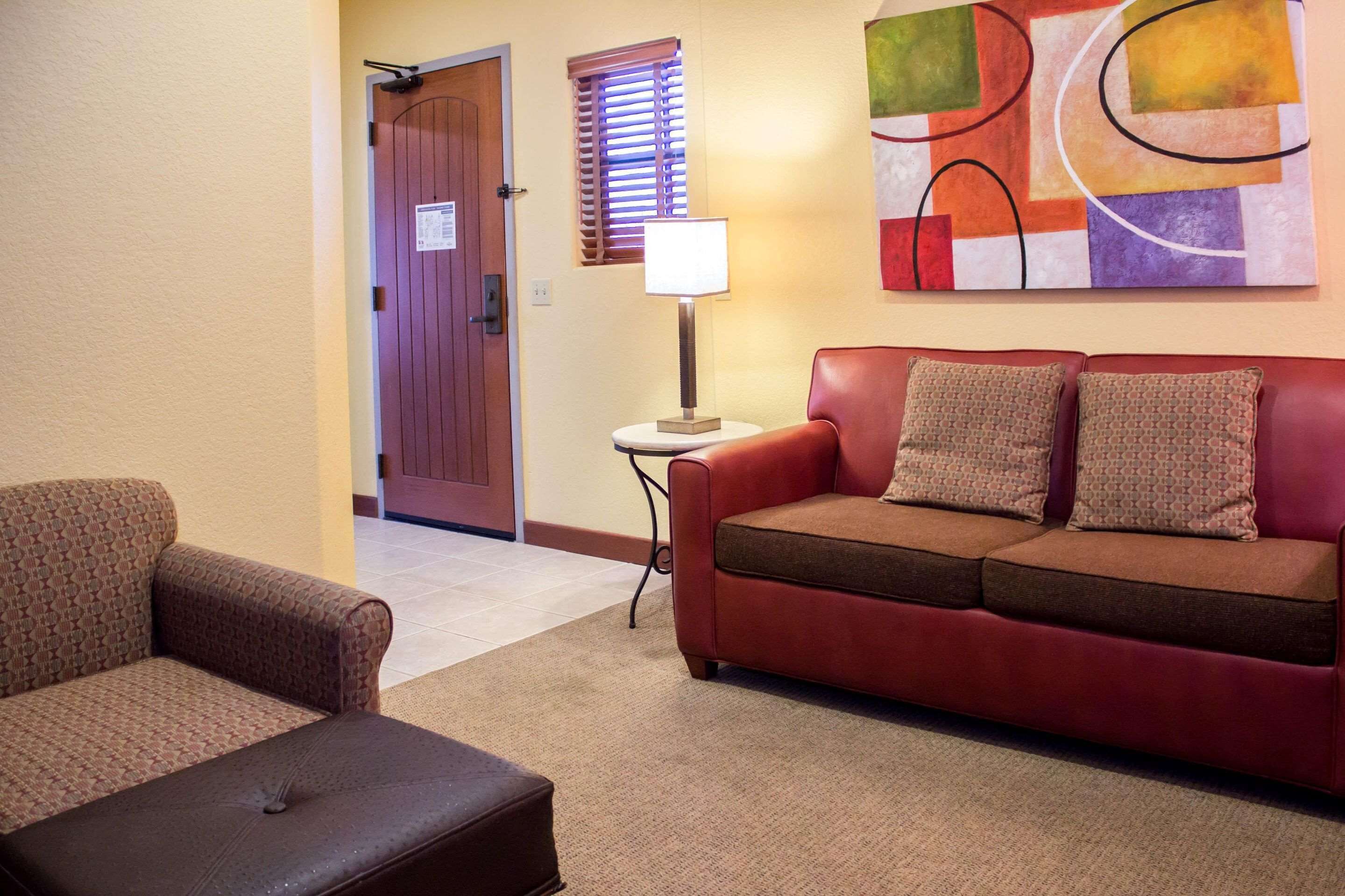 A hotel room with a red leather sofa, a patterned armchair, a small table with a lamp, and a door with a sign.