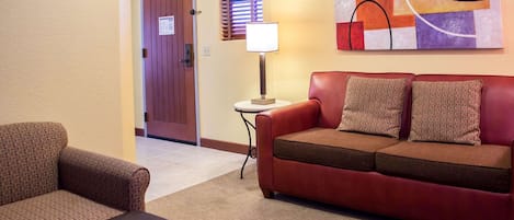 A hotel room with a red leather sofa, a patterned armchair, a small table with a lamp, and a door with a sign.