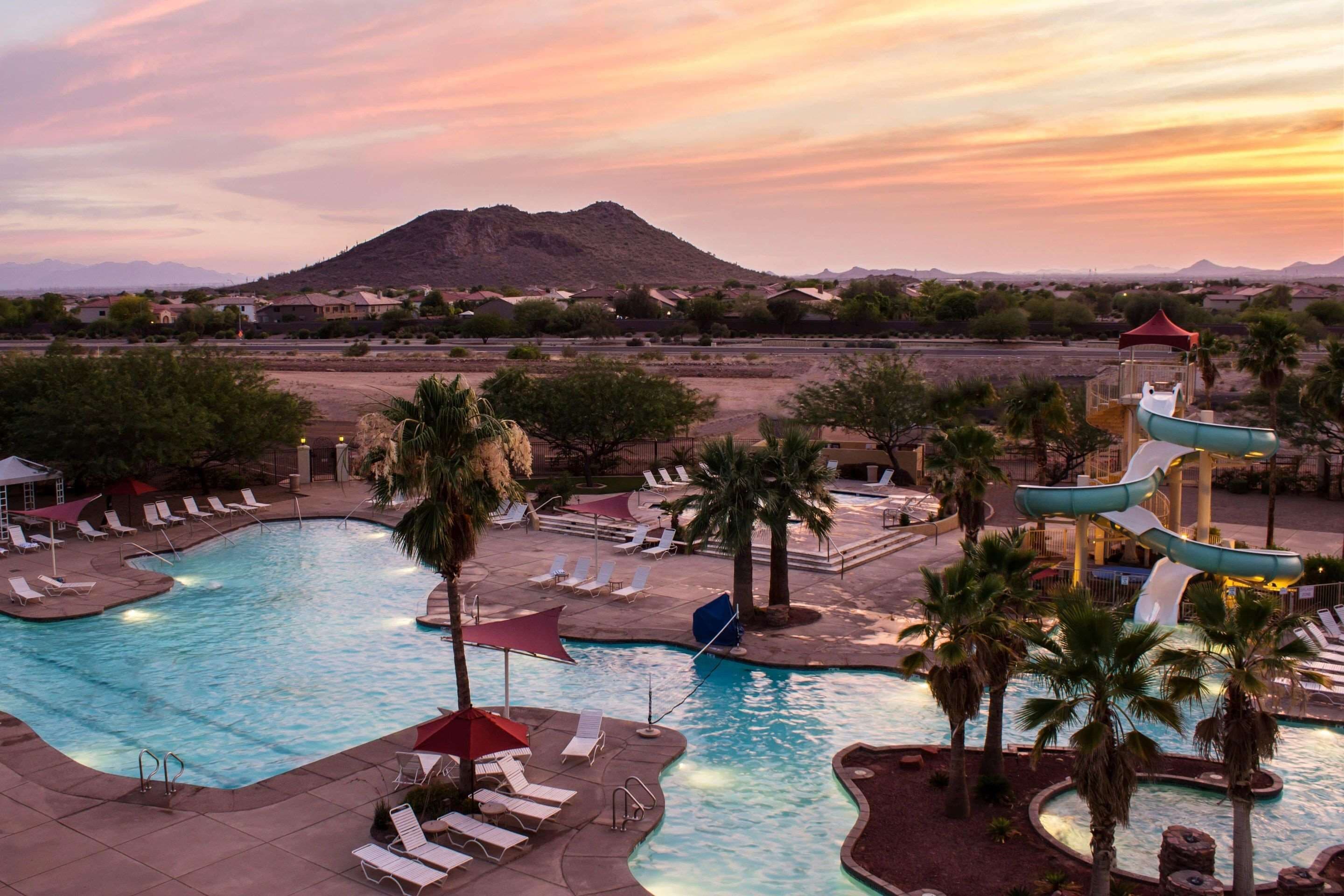 A swimming pool with lounge chairs and a water slide, surrounded by palm trees and a mountain in the background.