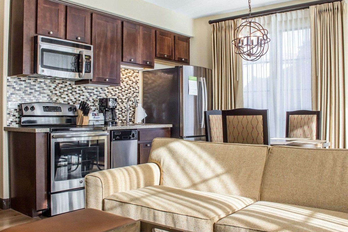 A compact living space with a kitchenette, including a microwave, stove, and refrigerator. There is a sofa and a dining area with chairs.
