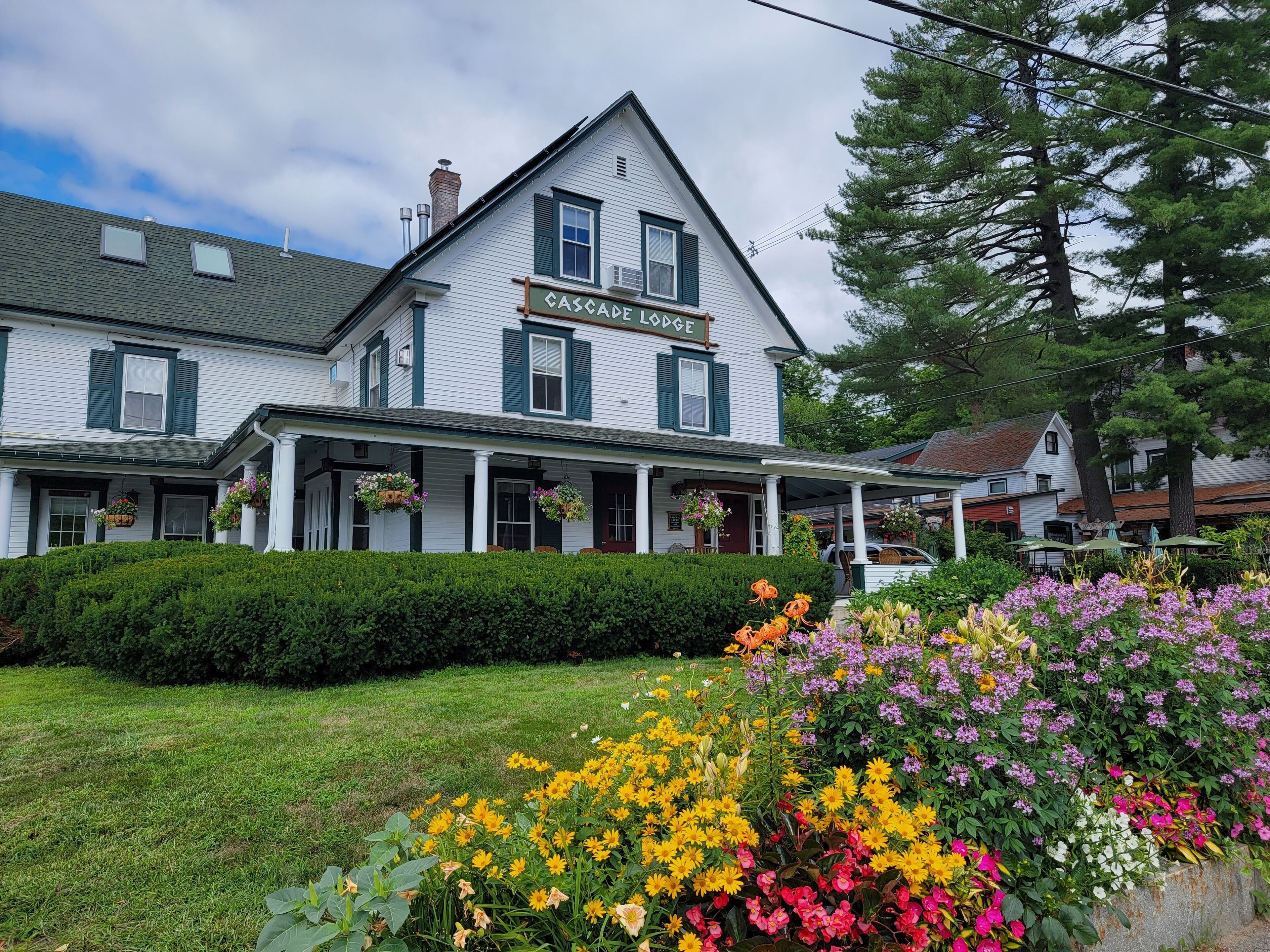 A two-story building with a sign reading "Cascade Lodge." It features a covered porch with hanging flower baskets and a well-maintained lawn with colorful flowers.