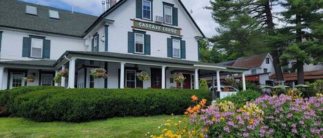A two-story building with a sign reading "Cascade Lodge." It features a covered porch with hanging flower baskets and a well-maintained lawn with colorful flowers.