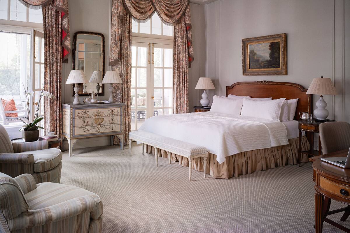 A classic hotel room with a large bed, two bedside tables with lamps, a dresser, a chair, and a window with curtains.