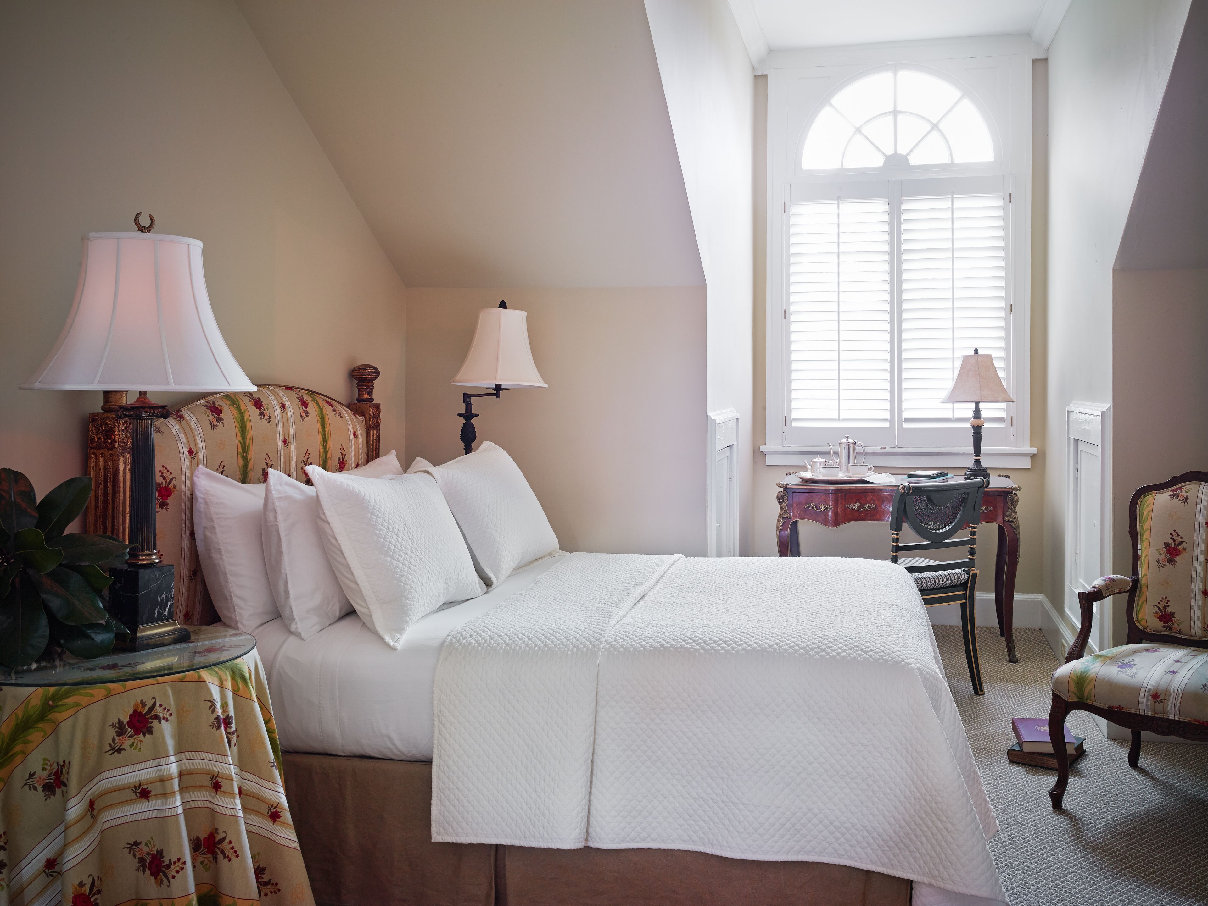 A bedroom with a large bed, two bedside lamps, a small table with a lamp, a chair, and a window with blinds.